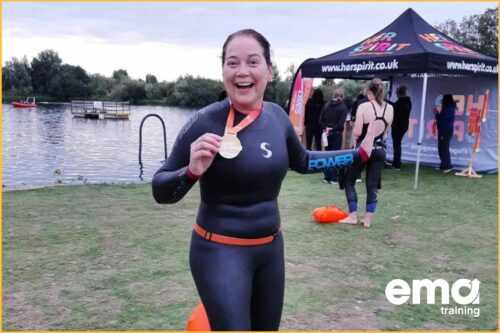 EMA's Mental Health First Aider, Julie holding up a medal in a wetsuit, smiling, after doing an open water charity swim.