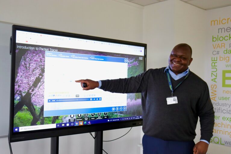 EMA's Digital Trainer, Admire, teaching an IT Apprenticeship class. Admire is standing in front of a big digital screen, pointing to the content on there.