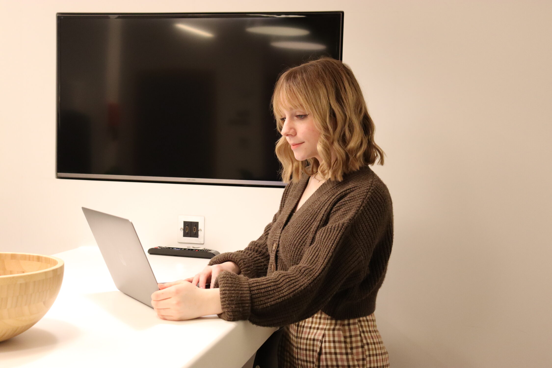 Digital Support Apprentice, Ruby. Ruby is pictured at a standing desk, working on a laptop, wearing a brown cardigan and checked trousers.