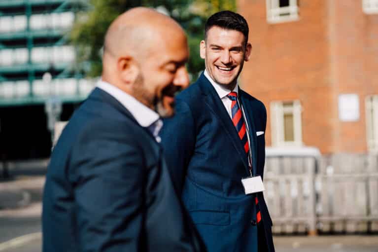 James, EMA's COO, smiling at a colleague. Both are in blue suits, standing outside of the EMA Training Finance Hub.