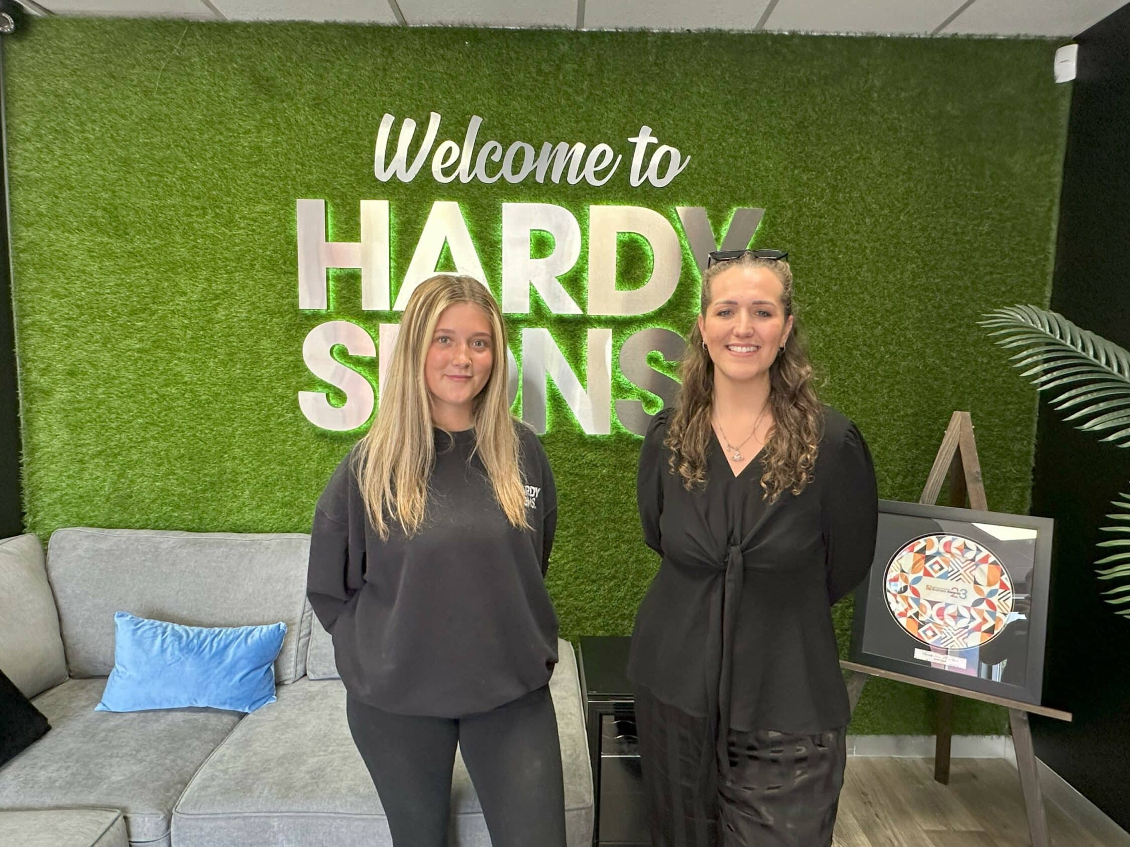 Blu and Chloe standing in front of a green plant wall with a large silver Hardy Signs sign